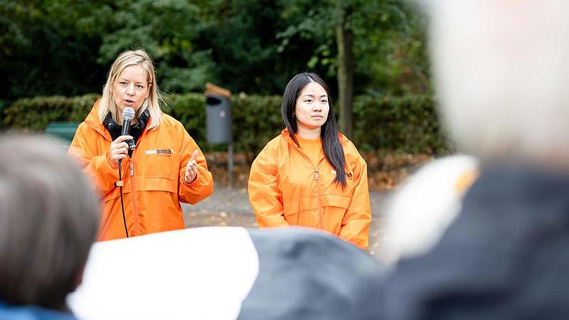 Zwei foodwatch Aktivistinnen in orangen Jacken sprechen mit einem Mikrofon zu den Demonstranten während des Protests. Sie stehen im Freien vor Bäumen.