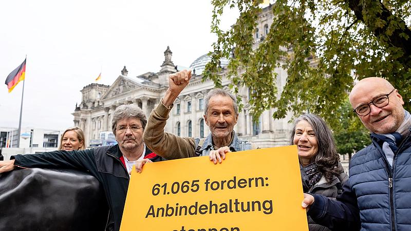 Eine Gruppe von Aktivisten steht vor dem deutschen Bundestag mit einem großen gelben Plakat. Auf dem Plakat steht: "61.065 fordern: Anbindehaltung stoppen." Ein Mann hebt seine Faust, um den Protest zu unterstützen.