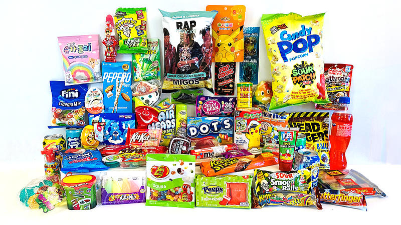 Große Ansammlung bunt verpackter Süßigkeiten auf weißem Hintergrund. Zu sehen sind Produkte wie „Sour Patch“, „Warheads“, „Pop Rocks“ und „Candy Pop Popcorn“. Viele Verpackungen nutzen grelle Farben und Comic-Figuren. Auch Energy-Drinks und saure Gele sind dabei.