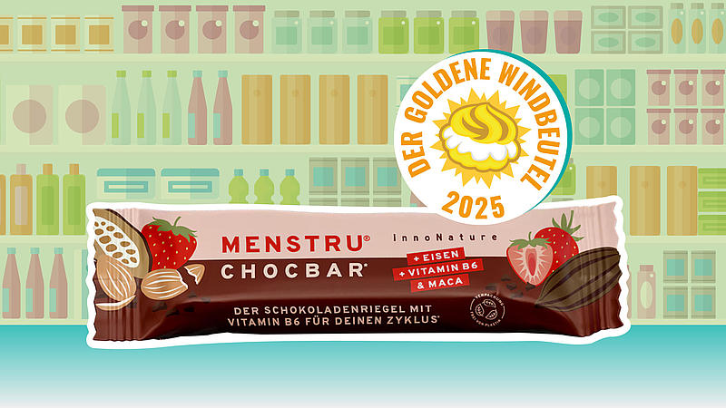 Quer liegende Verpackung des Schokoladenriegels „Menstru Chocbar“ (Erdbeere, Kakaobohne-Grafiken) vor Regal-Illustration; rechts oben das Siegel „Der Goldene Windbeutel 2025“.