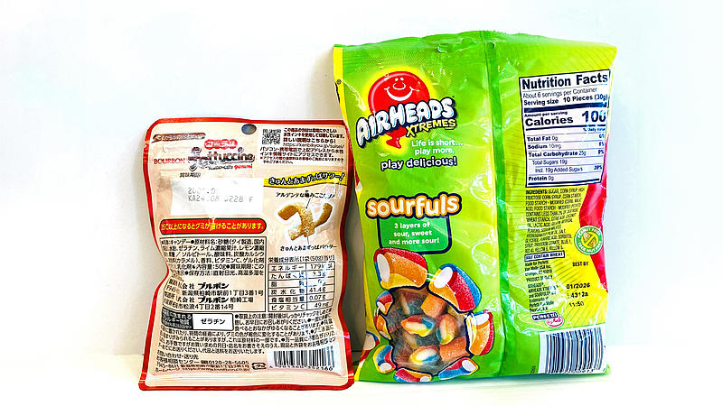 Zwei Rückseiten von Süßigkeitenverpackungen. Links ein japanisches Produkt mit ausschließlich japanischer Beschriftung, rechts eine Packung „Airheads Xtremes Sourfuls“ mit farbenfrohem Design und englischer Nährwerttabelle. Zu erkennen sind viele Zuckerarten und künstliche Zusätze.