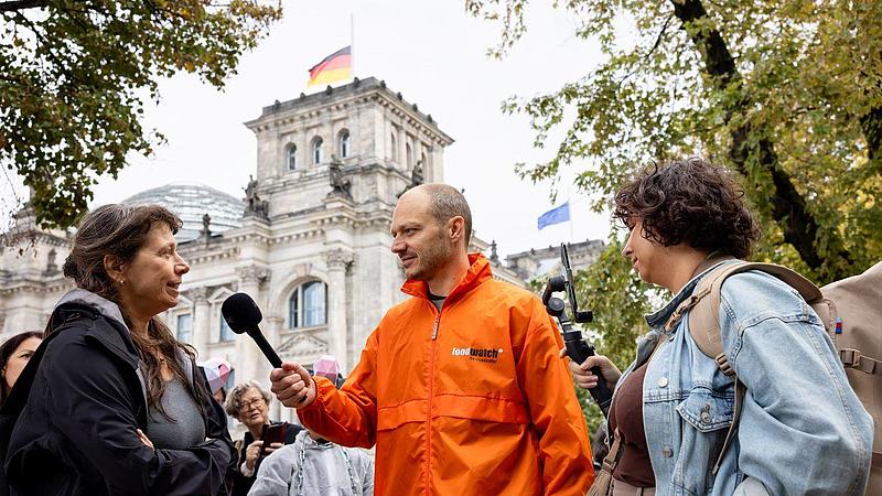 Ein Aktivist in einer orangen Foodwatch-Jacke interviewt eine Frau vor dem deutschen Bundestag. Im Hintergrund sind weitere Demonstranten zu sehen.
